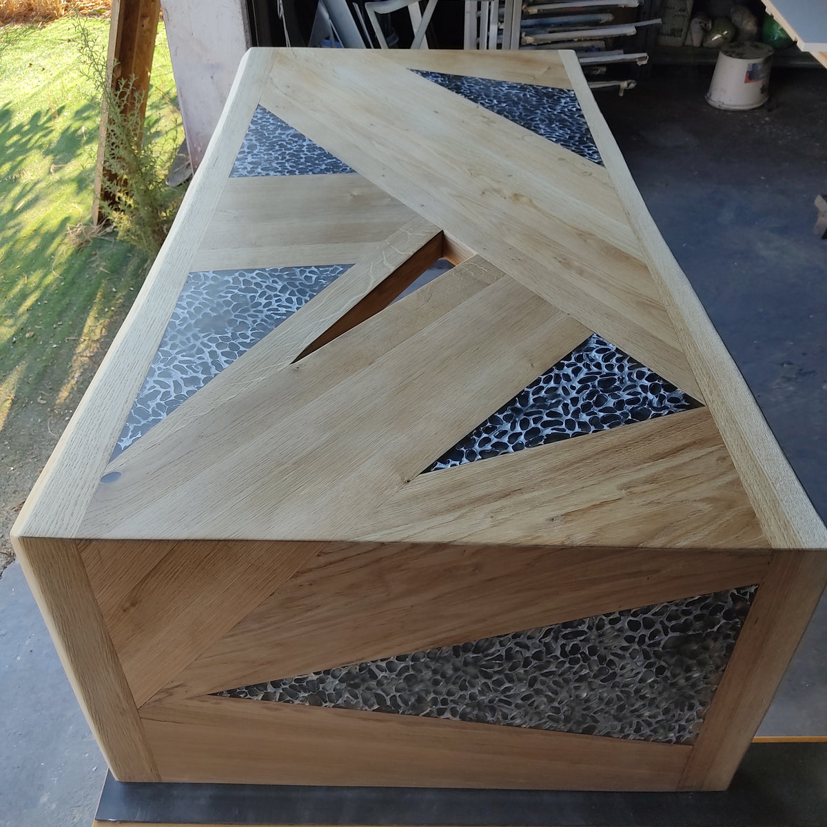tavolo in quercia, superficie in rovere massello sagomata in pannelli geometrici, con inserti a triangoli di resina traslucida a effetto ciottolato. Linee pulite e volumi decisi definiscono un’estetica contemporanea.