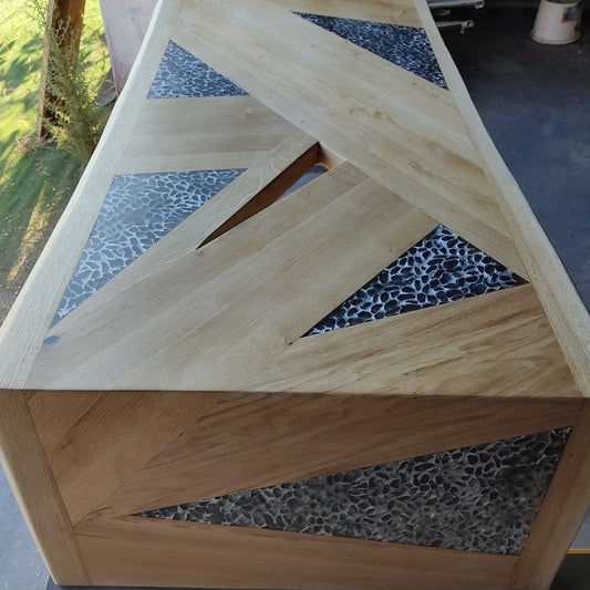 tavolo in quercia, superficie in rovere massello sagomata in pannelli geometrici, con inserti a triangoli di resina traslucida a effetto ciottolato. Linee pulite e volumi decisi definiscono un’estetica contemporanea.
